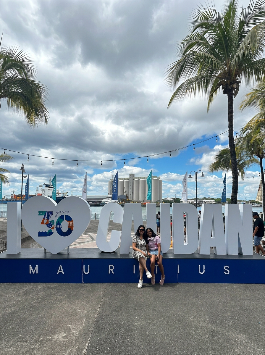IMG_1469 Two women sitting in front of a large sign that reads 'I LOVE CAUDAN' celebrating 30 years in Mauritius, with a cloudy sky and palm trees in the background.