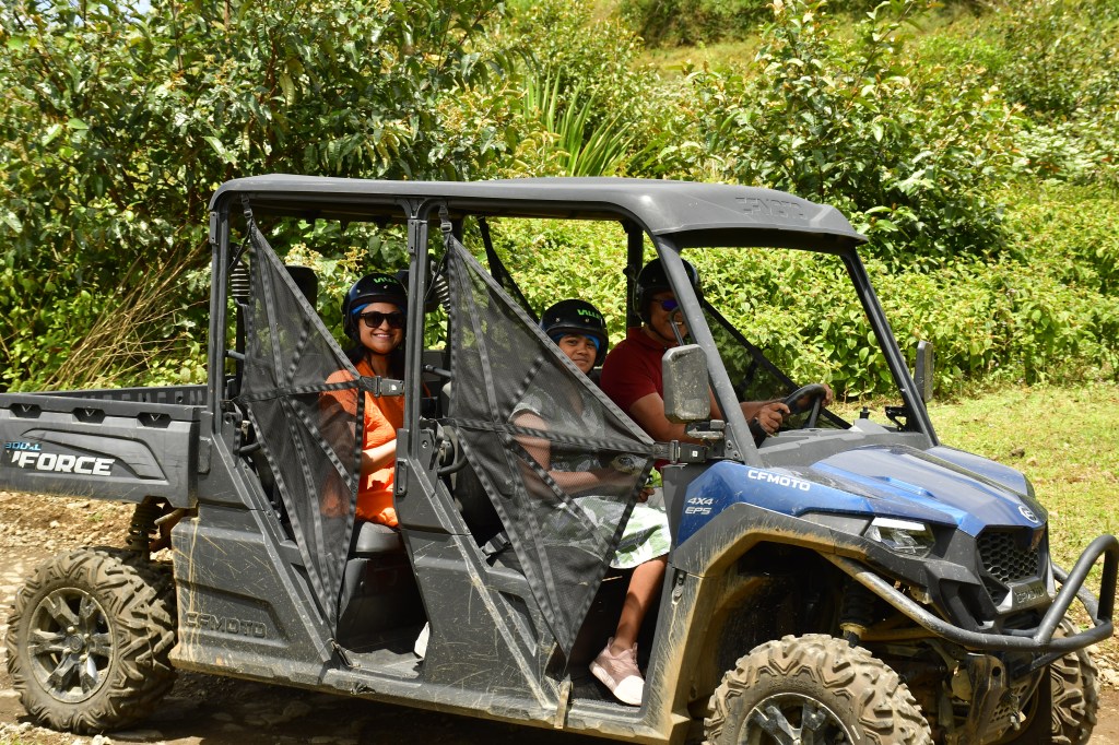 A group of three people, including two wearing helmets, are sitting inside a utility vehicle on a sunny day, surrounded by lush greenery.