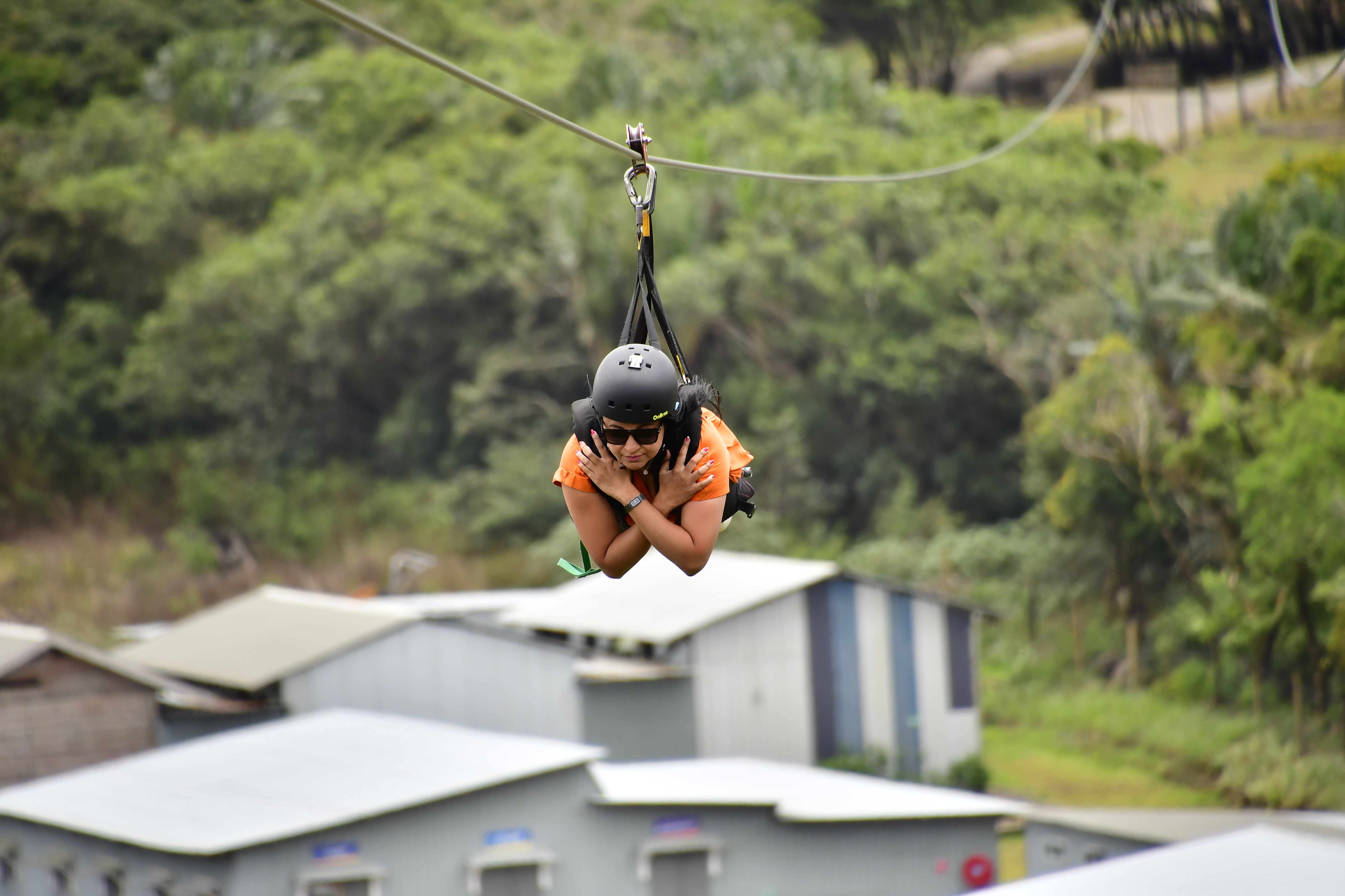 A person zip-lining in an orange shirt and helmet, suspended above a landscape with green trees and gray buildings in the background.