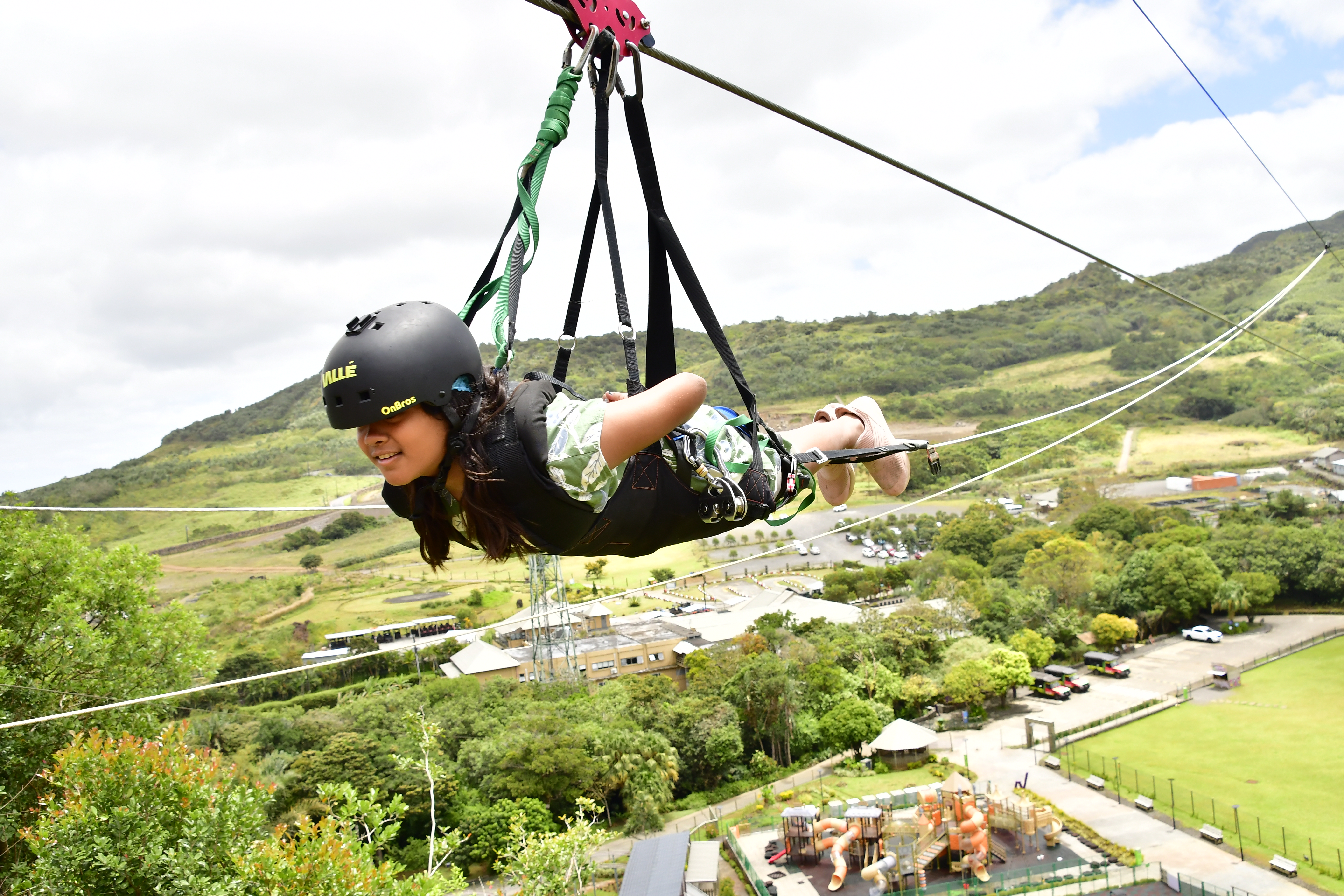 A young girl with a helmet gliding through the air on a zip line, surrounded by green hills and a playground below.