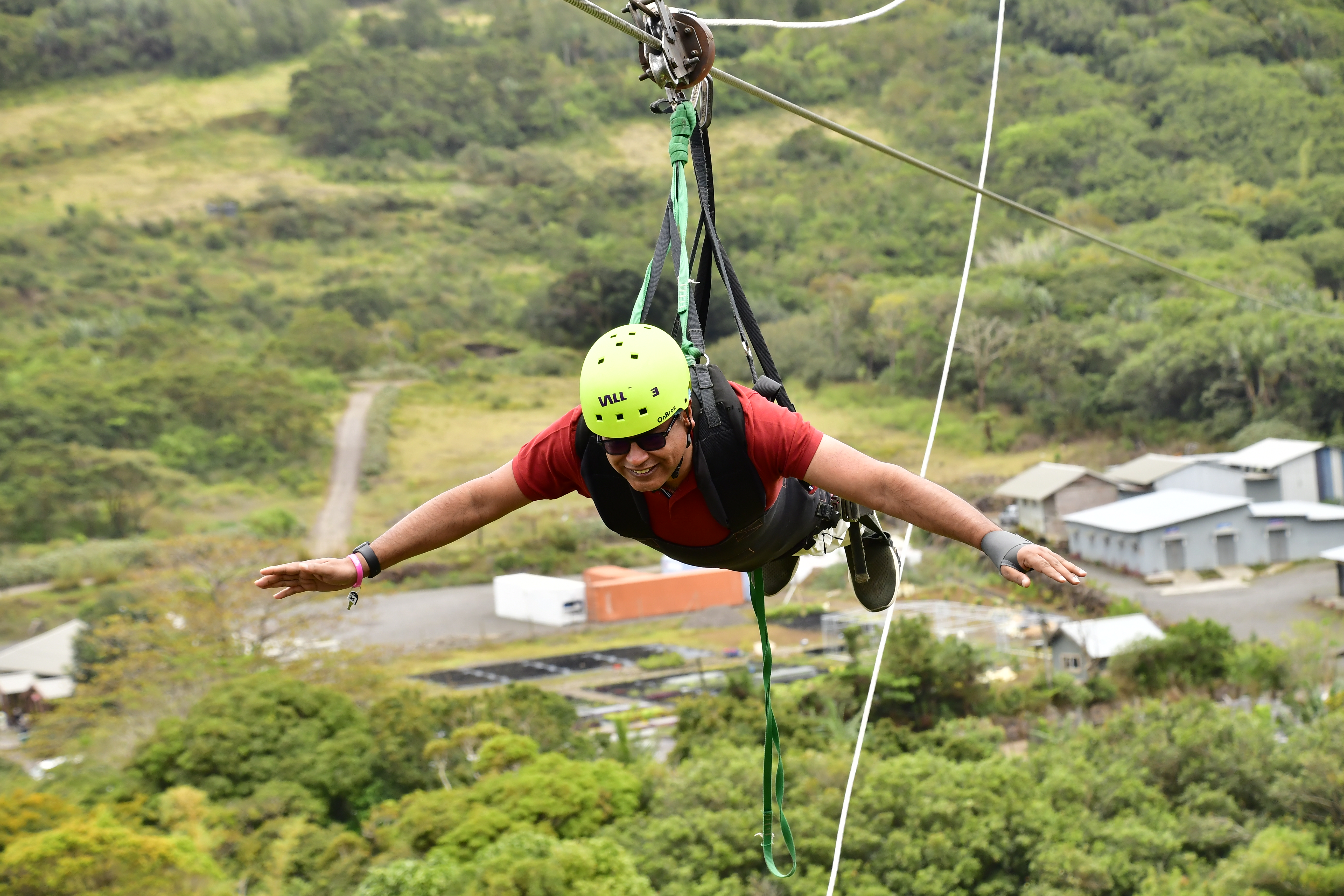 A person enjoying a zipline ride, arms extended, wearing a green helmet and a red shirt, with a lush green landscape and buildings in the background.