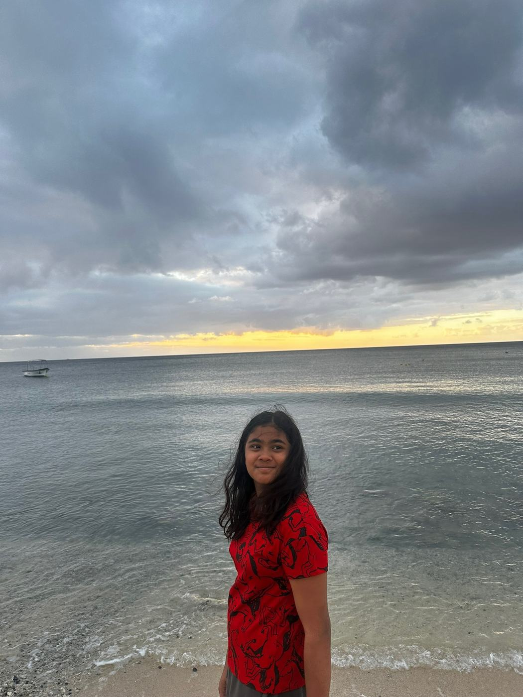 A girl with long hair stands on the beach, wearing a red shirt with a design, looking towards the sea as the sun sets, creating a picturesque view of clouds and water.