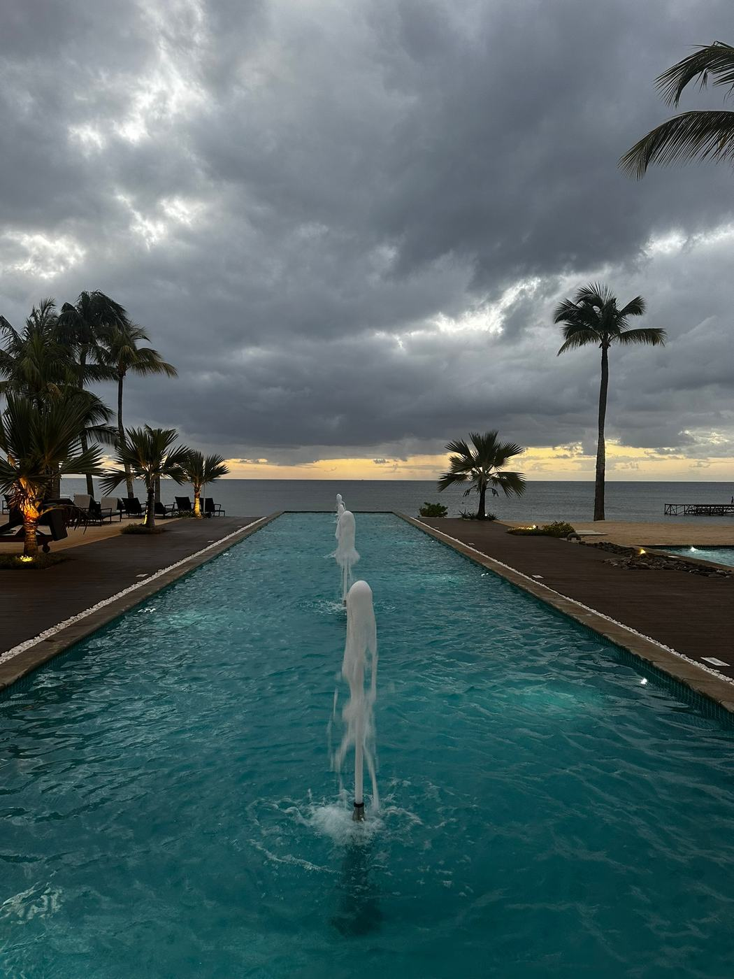 A tranquil resort pool featuring several water fountains, surrounded by palm trees, with a cloudy sky and ocean in the background during twilight.