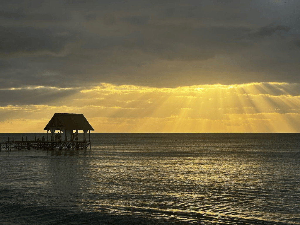 A serene sunset over the ocean with rays of sunlight breaking through the clouds, illuminating a wooden pier with a thatched roof.