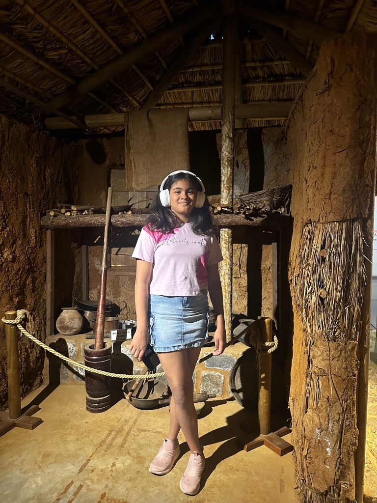 A young girl wearing headphones stands inside a rustic, traditional dwelling made of mud and thatch, surrounded by historical artifacts.
