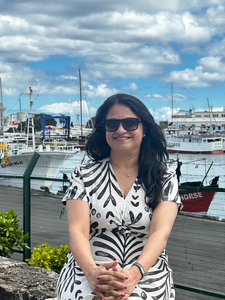A woman sitting by a harbor, wearing sunglasses and a patterned dress, with boats and a cloudy sky in the background.