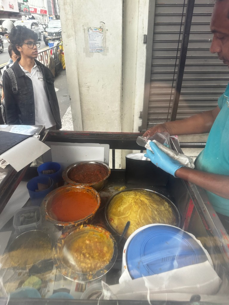 A street food vendor preparing dishes with various colorful curries and lentils in bowls, while a customer watches in the background.