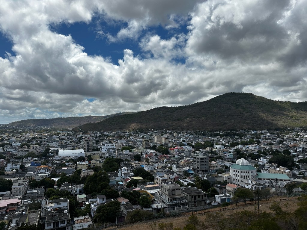 A panoramic view of a city with various buildings, including residential and commercial structures, situated at the base of a hilly landscape under a cloudy sky.