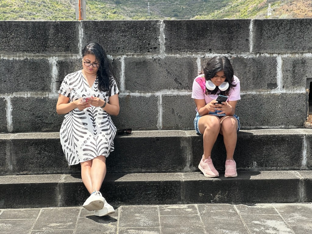 Two young women sitting on stone steps, both looking at their mobile phones. One is wearing a patterned dress and glasses, while the other is in a pink shirt with headphones around her neck.