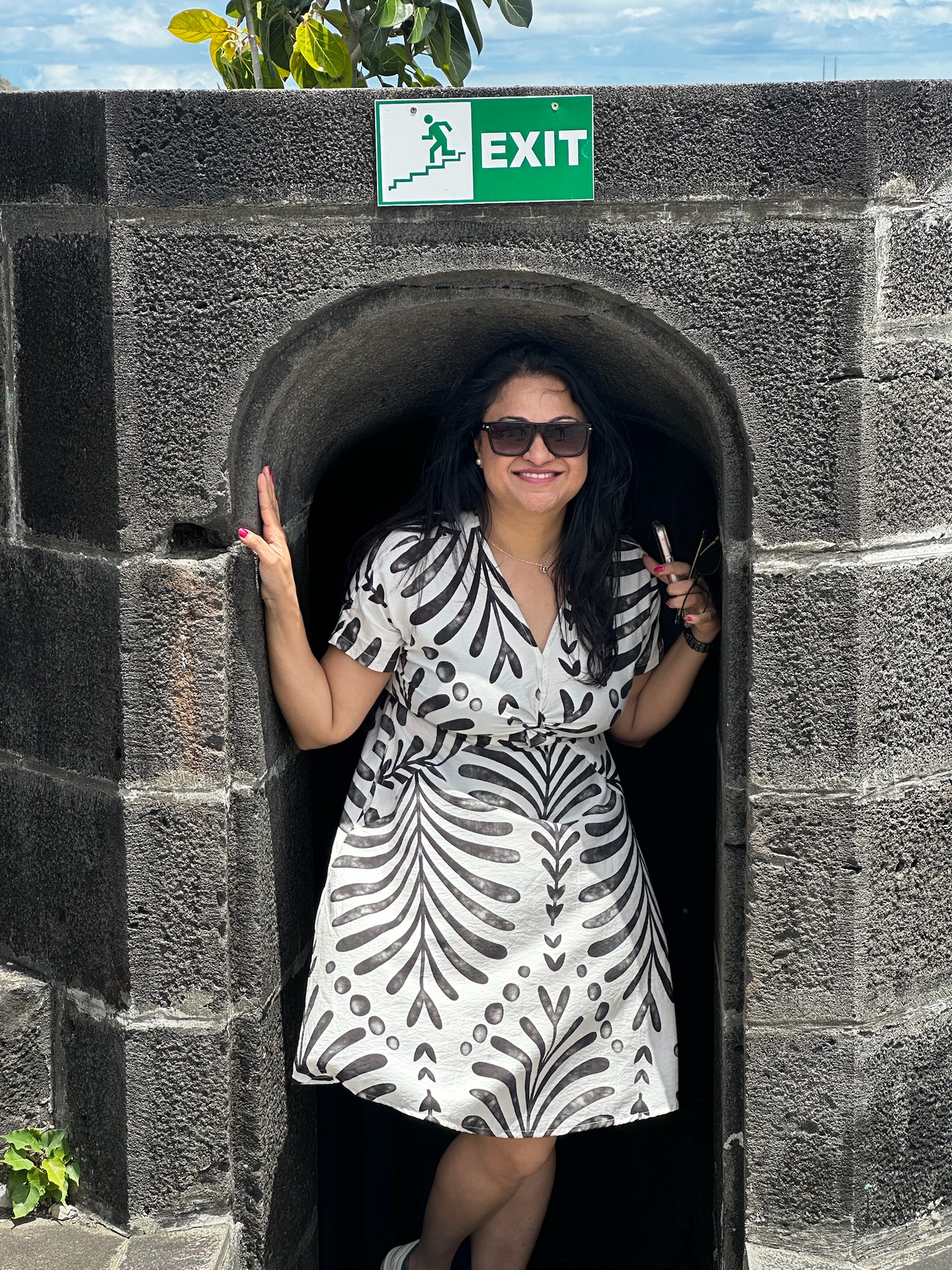 A woman standing in a stone archway with a sign above that says 'EXIT,' wearing sunglasses and a patterned dress.