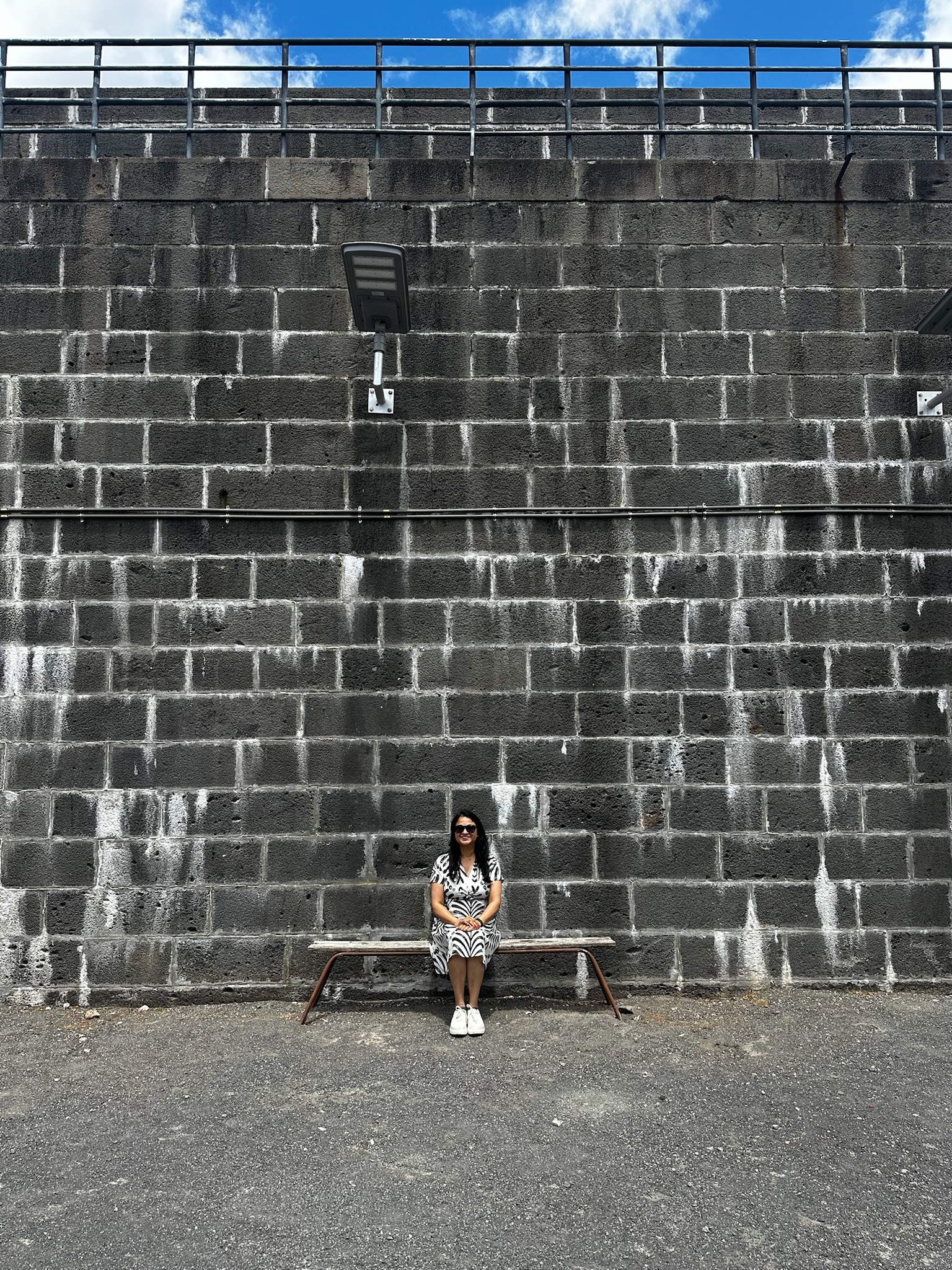 A person sitting on a bench in front of a textured gray wall with a light fixture above and blue sky with clouds in the background.