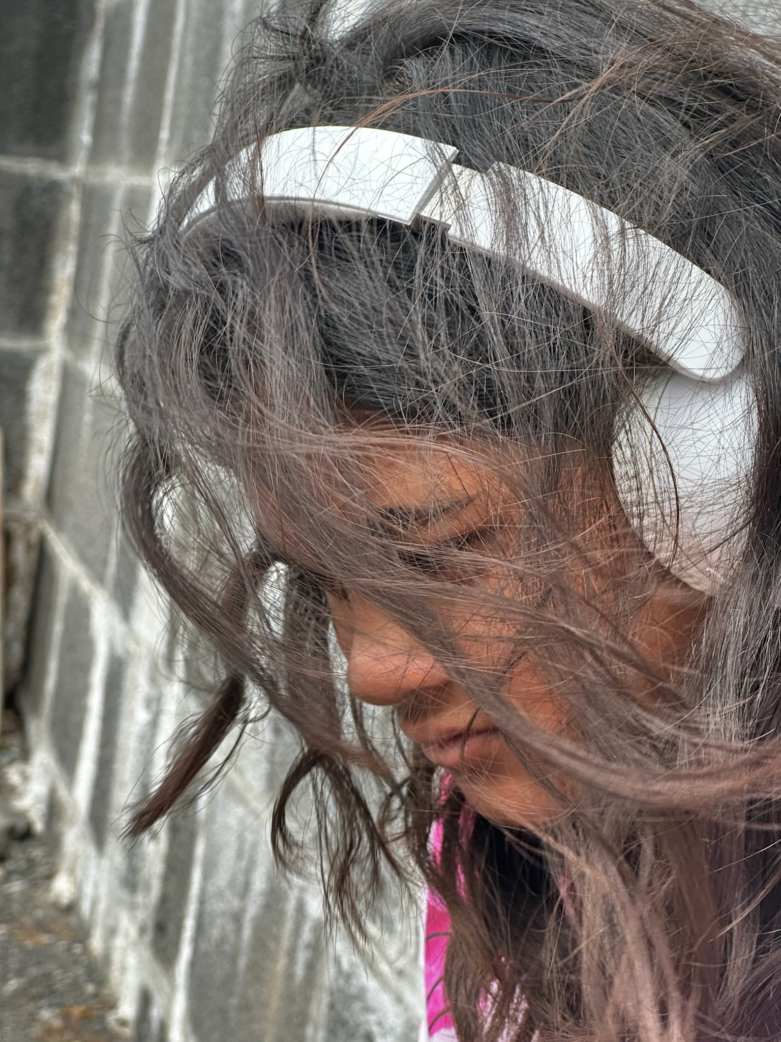 A young person with long, windblown hair is wearing white headphones and looking down. The background features a textured gray wall.