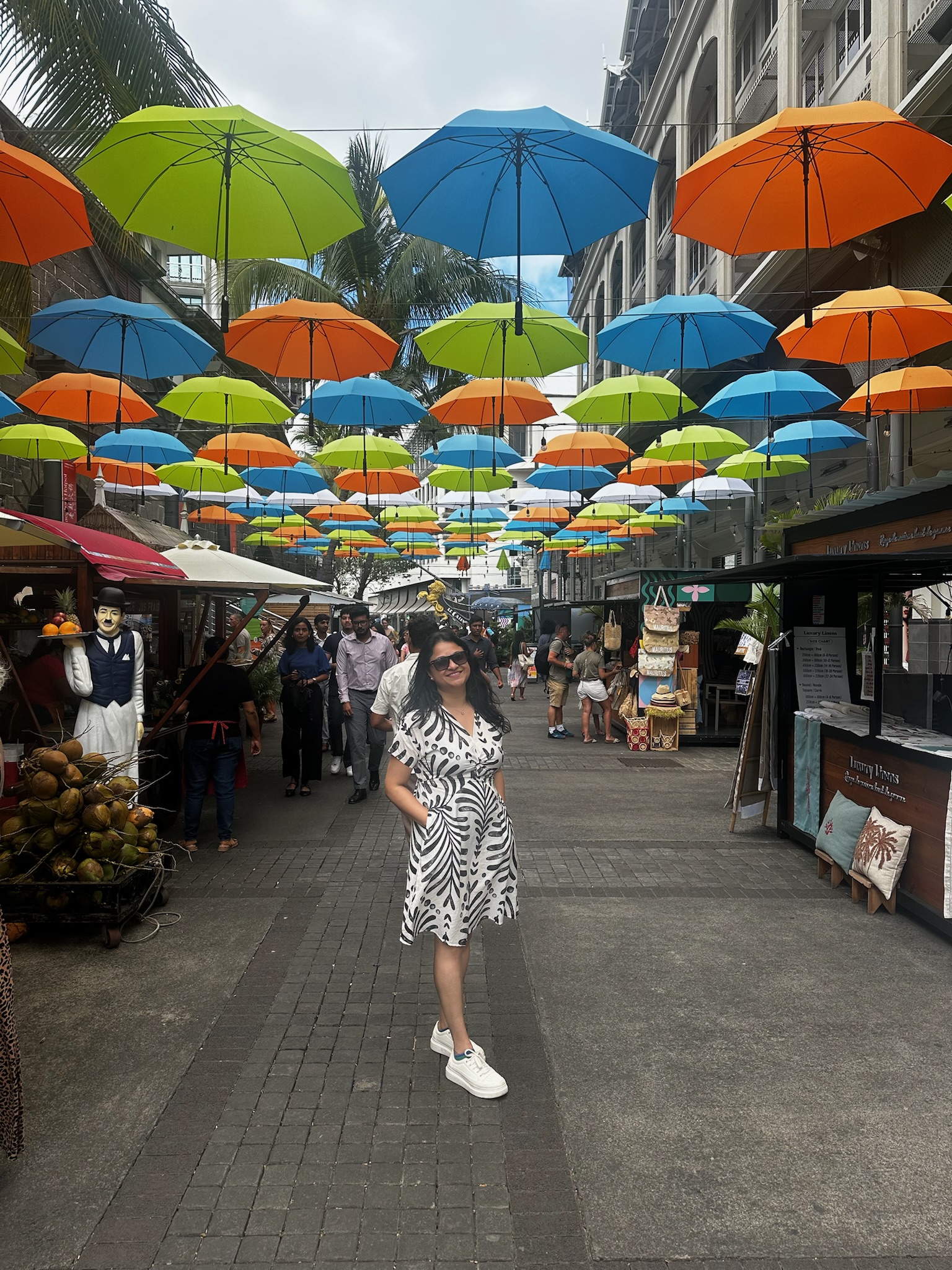 A woman in a black and white patterned dress stands smiling on a bustling street decorated with colorful umbrellas overhead. Various stalls and people are visible in the background, creating a vibrant scene.