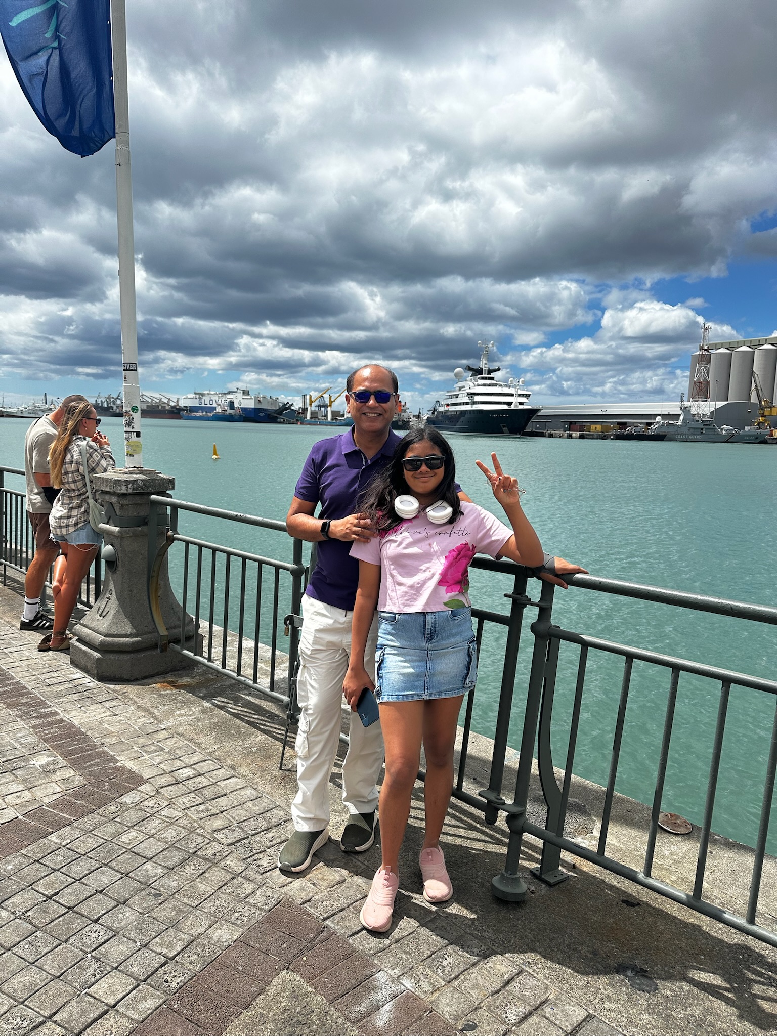 A man and a girl standing by a waterfront with a cloudy sky, smiling and posing for the photo. The girl is wearing a pink shirt and a denim skirt, showing a peace sign.