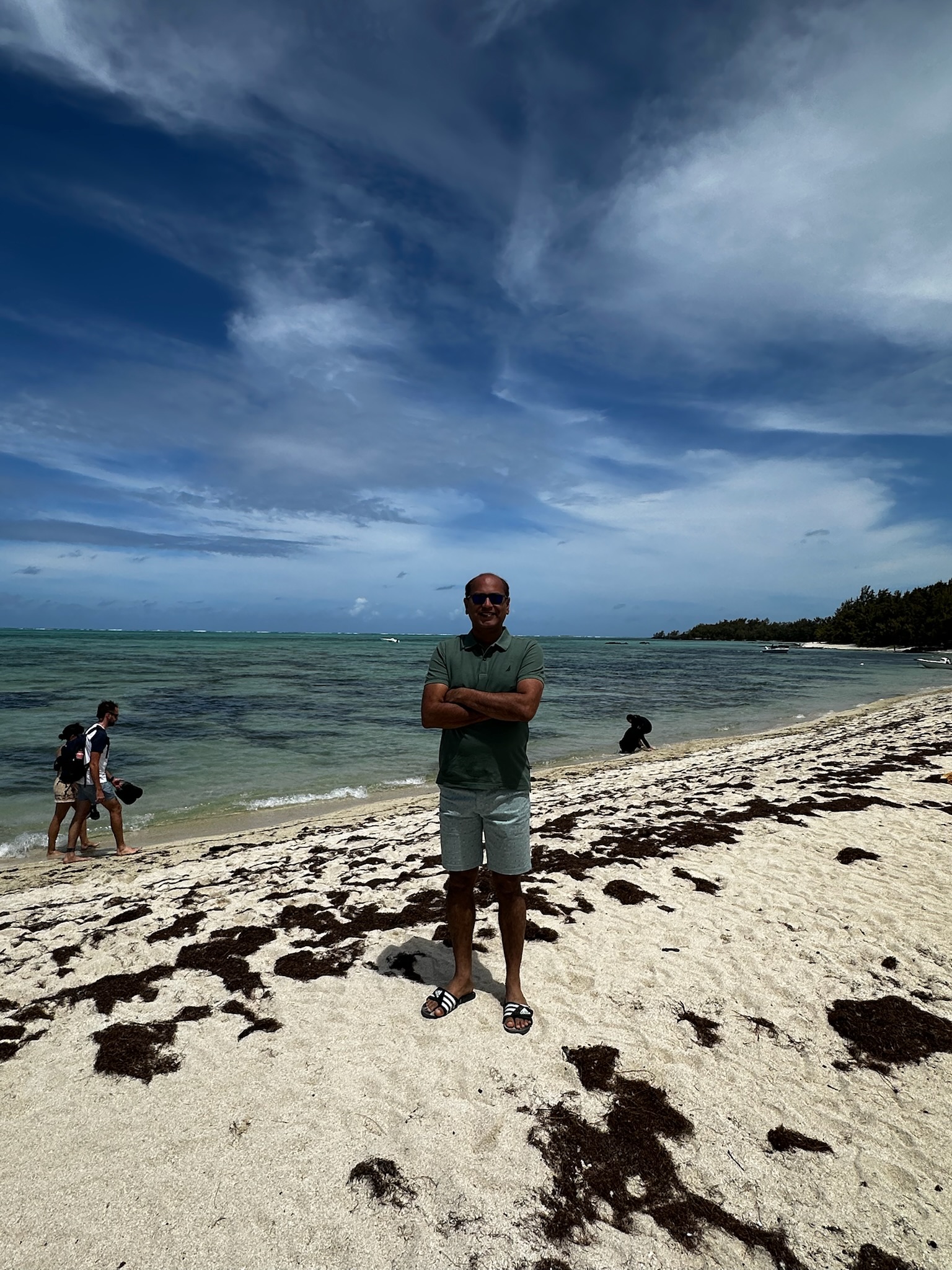 Person standing on a sandy beach with seaweed, wearing sunglasses and casual clothing, with a scenic ocean and cloudy sky in the background.