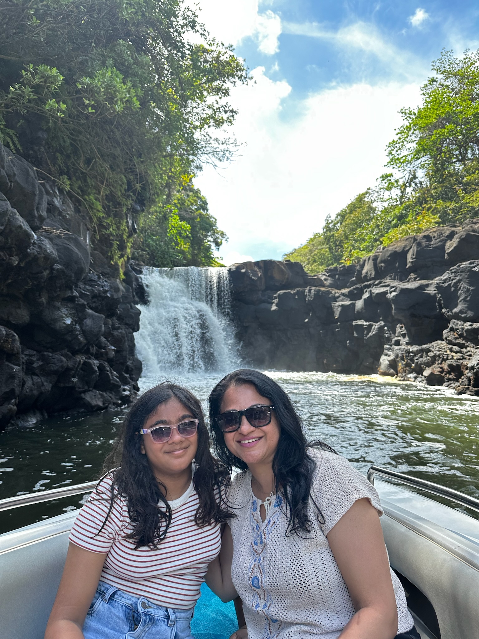 A mother and daughter smiling together on a boat near a waterfall, surrounded by lush greenery and rocky cliffs.
