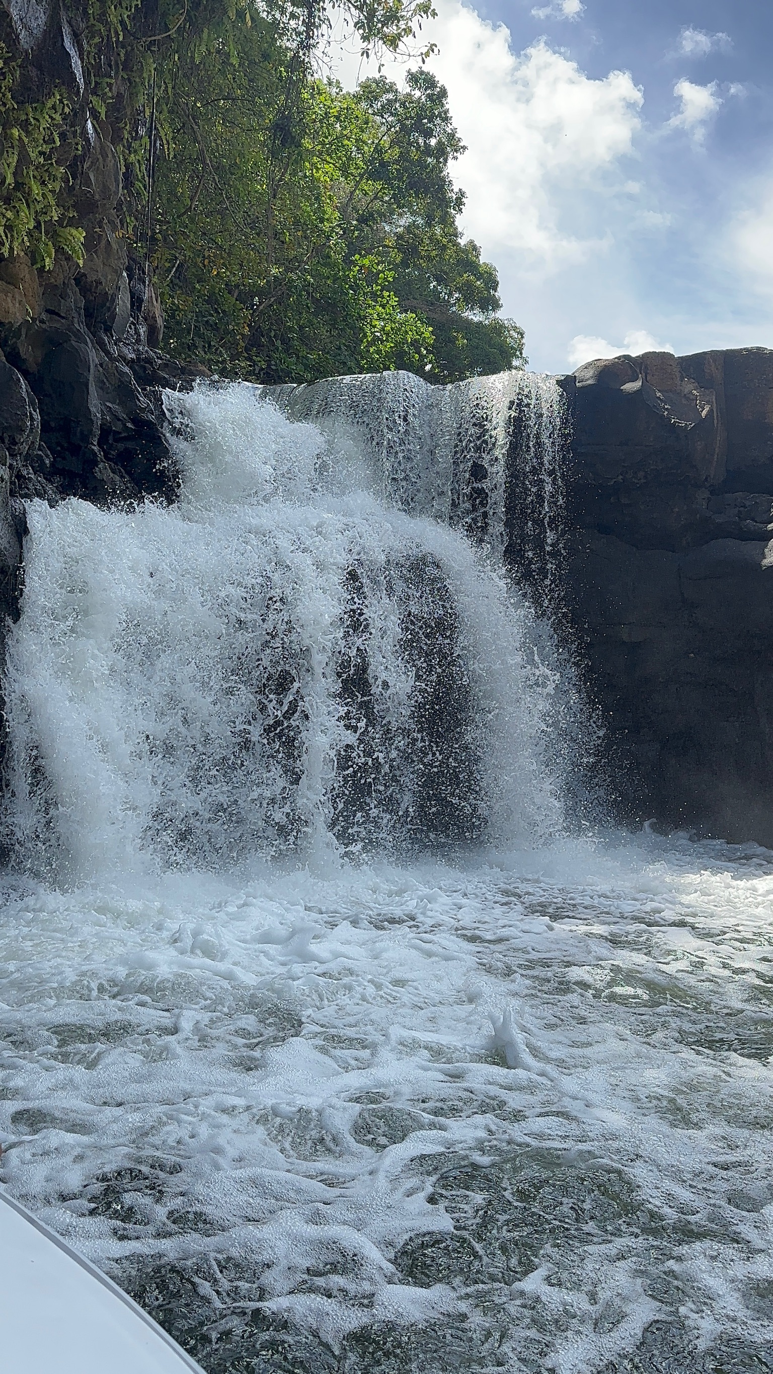 A picturesque waterfall cascading down rocky cliffs into a foamy pool below, surrounded by lush greenery and a clear blue sky.