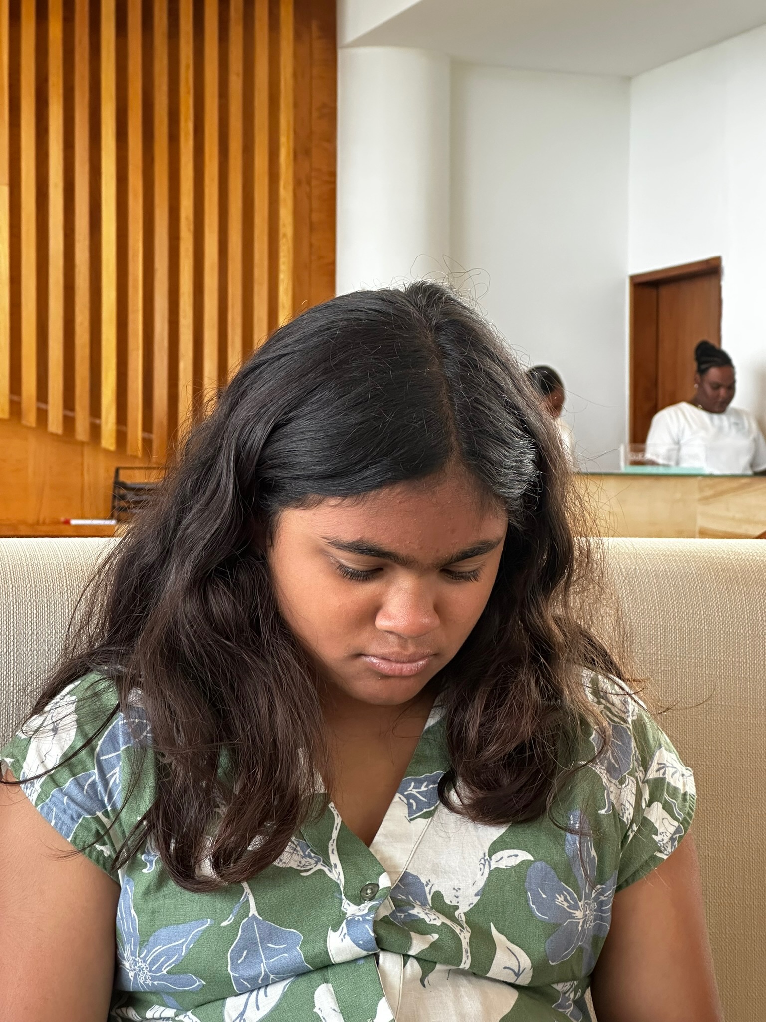 A girl with long, dark hair is sitting with her head down, wearing a floral green and white shirt. Soft wood paneling is visible in the background.