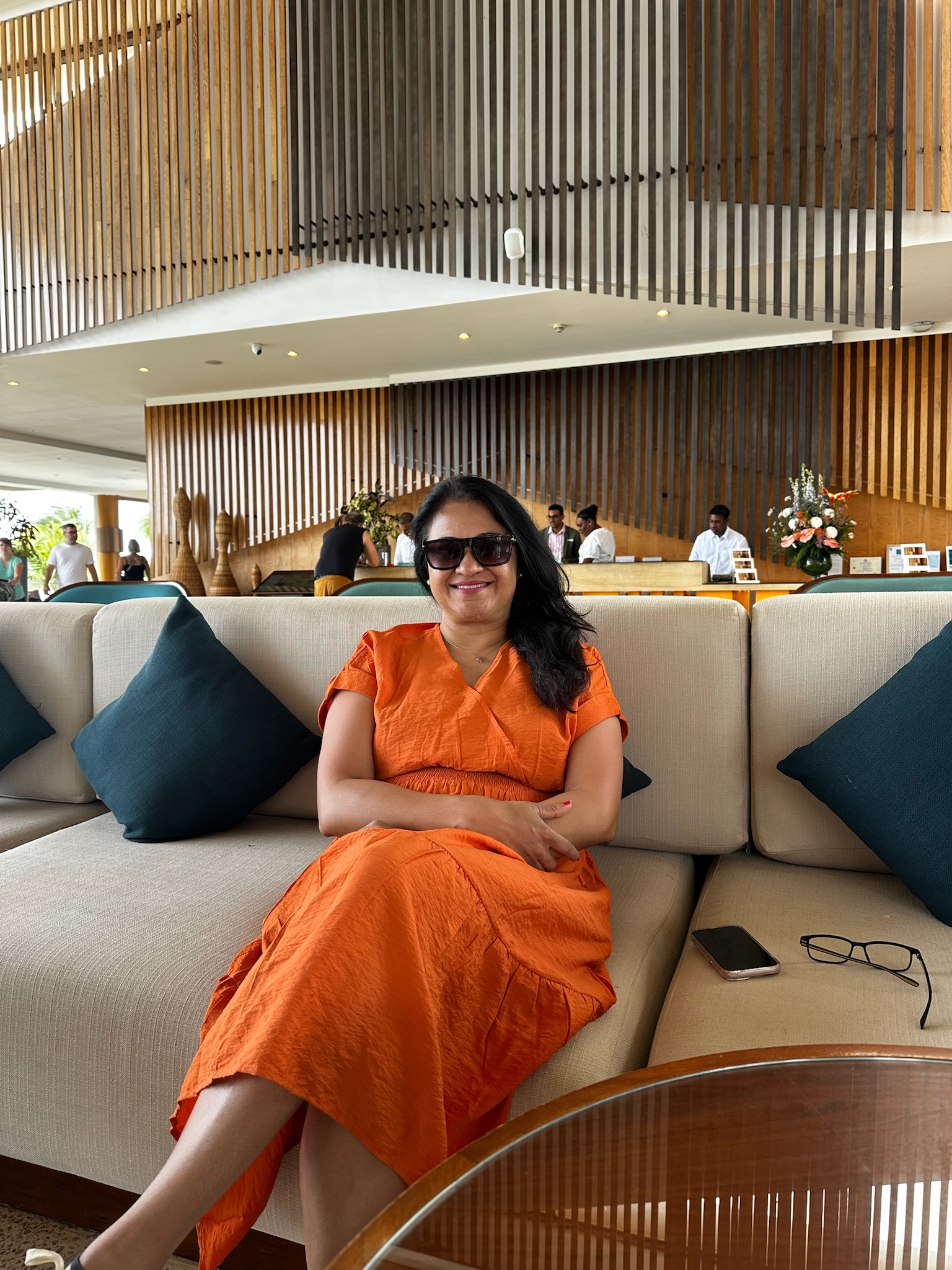 A woman in an orange dress sitting on a sofa with decorative cushions in a modern lobby, smiling and wearing sunglasses.