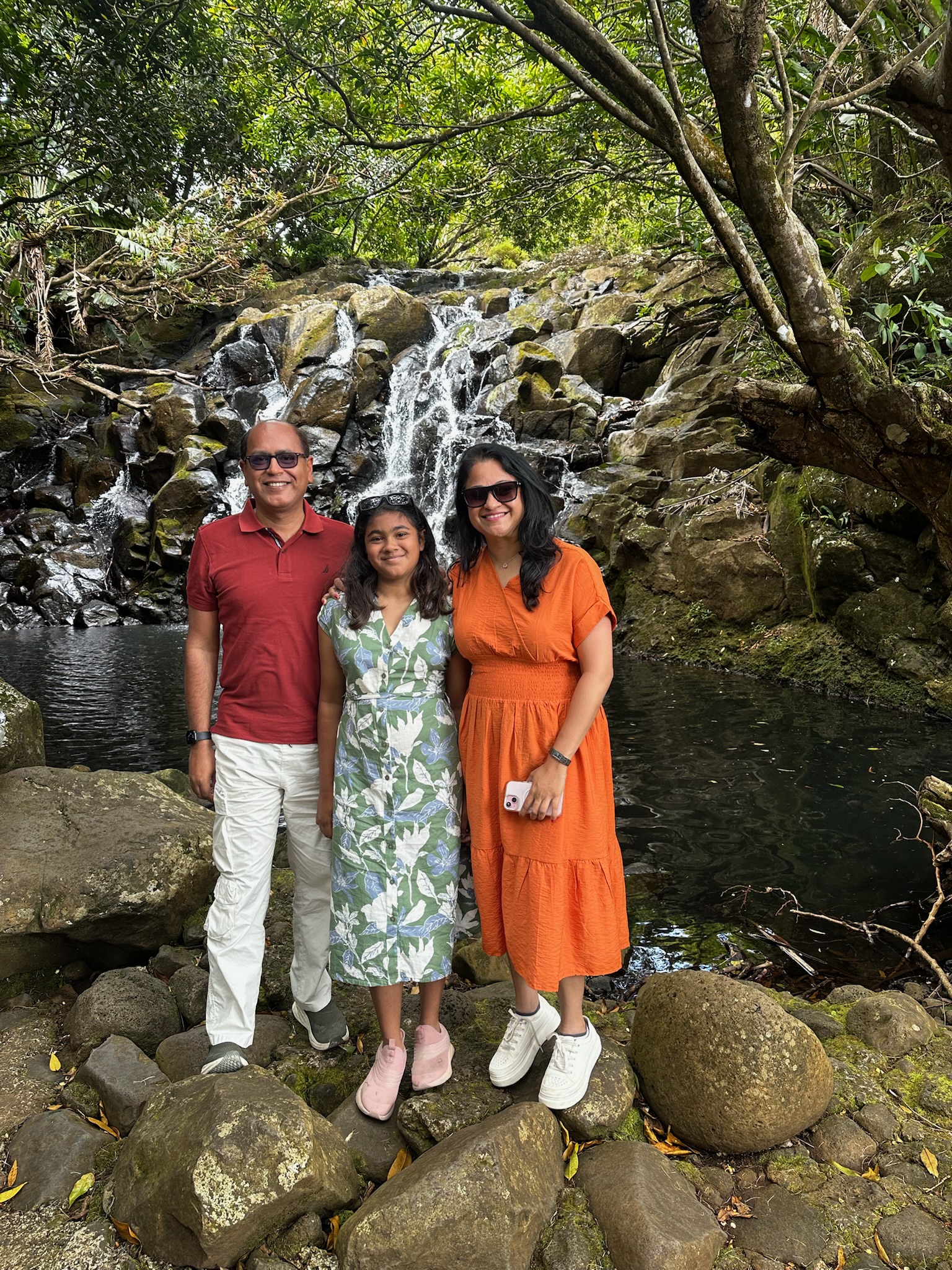 A family of three stands on rocks by a small waterfall in a lush, green environment. The father is wearing a red shirt and glasses, the daughter is in a floral dress, and the mother is in an orange dress. The background features rocks and flowing water.
