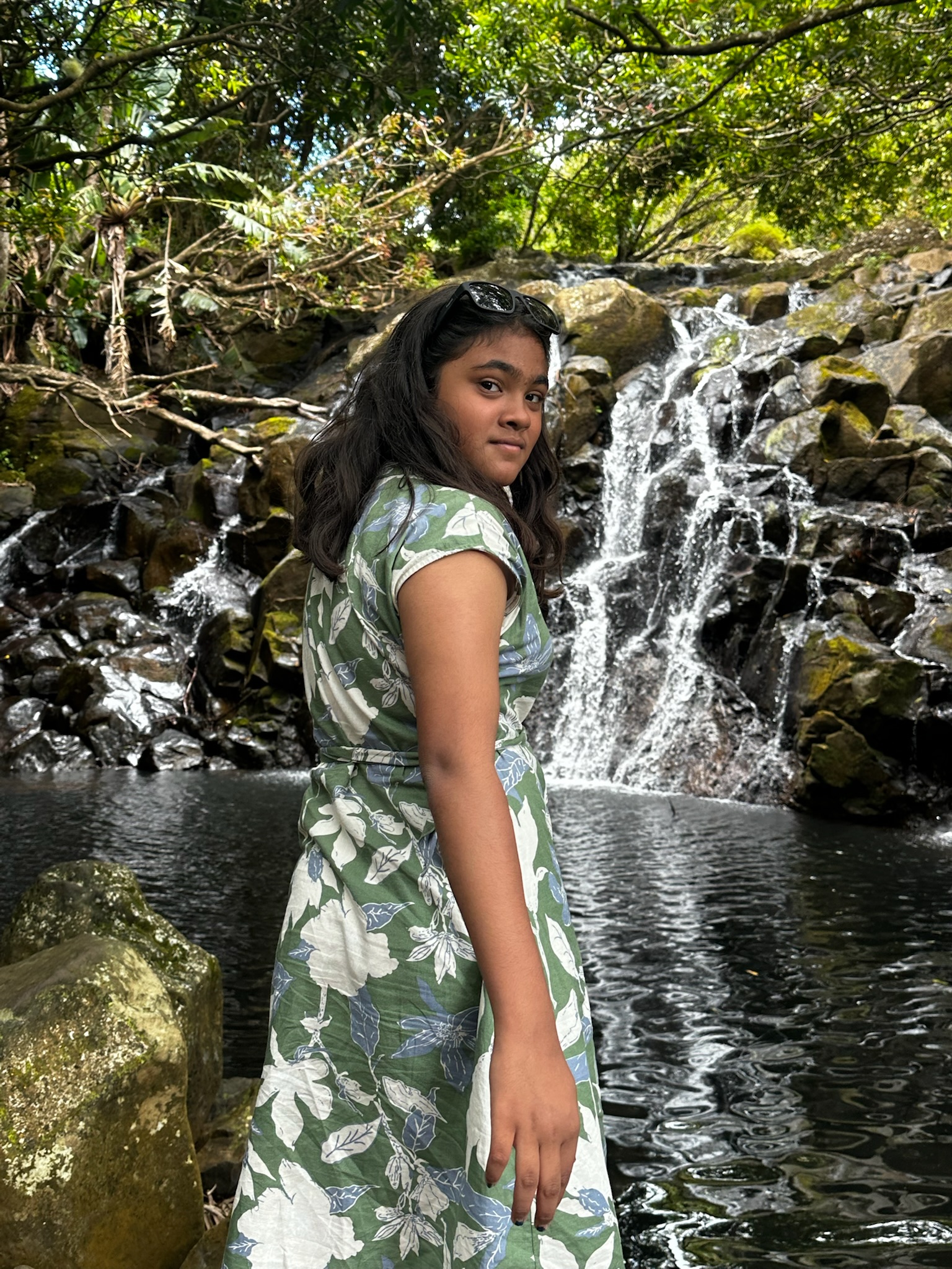 A girl with long dark hair, wearing a floral green dress, stands near a waterfall surrounded by lush greenery.