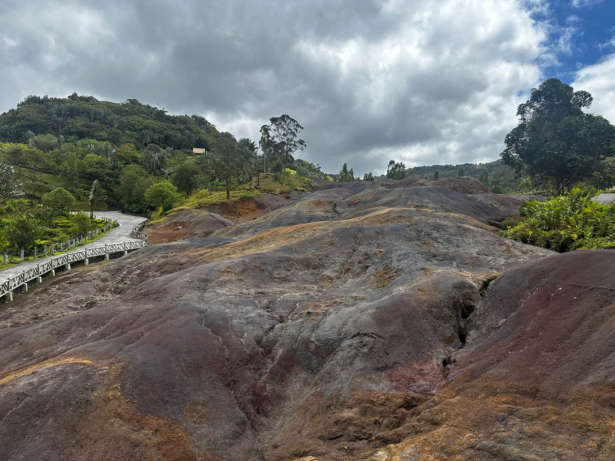 A landscape of colorful rocky terrain, with patches of brown, red, and gray rocks under a cloudy sky, and a winding path alongside lush greenery and trees.