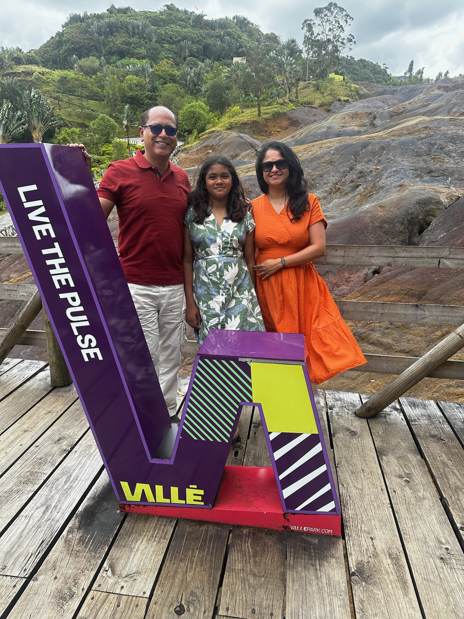 A family stands together in front of a colorful sign that says 'LIVE THE PULSE' and 'VALLĒ' on a wooden deck, with a scenic landscape of hills and greenery in the background.