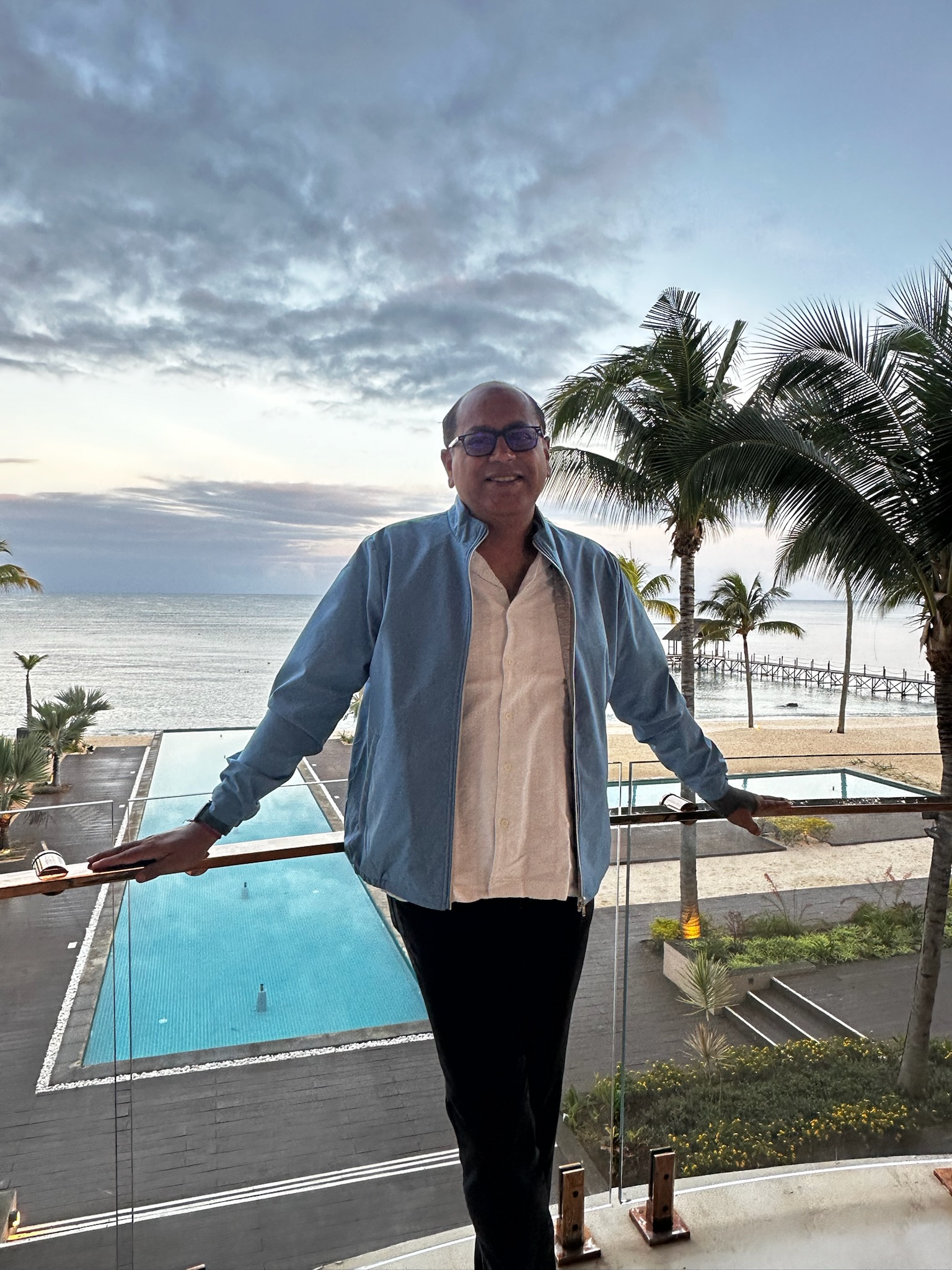 Man standing on a balcony overlooking a large swimming pool and the ocean, with palm trees in the background and a cloudy sky.