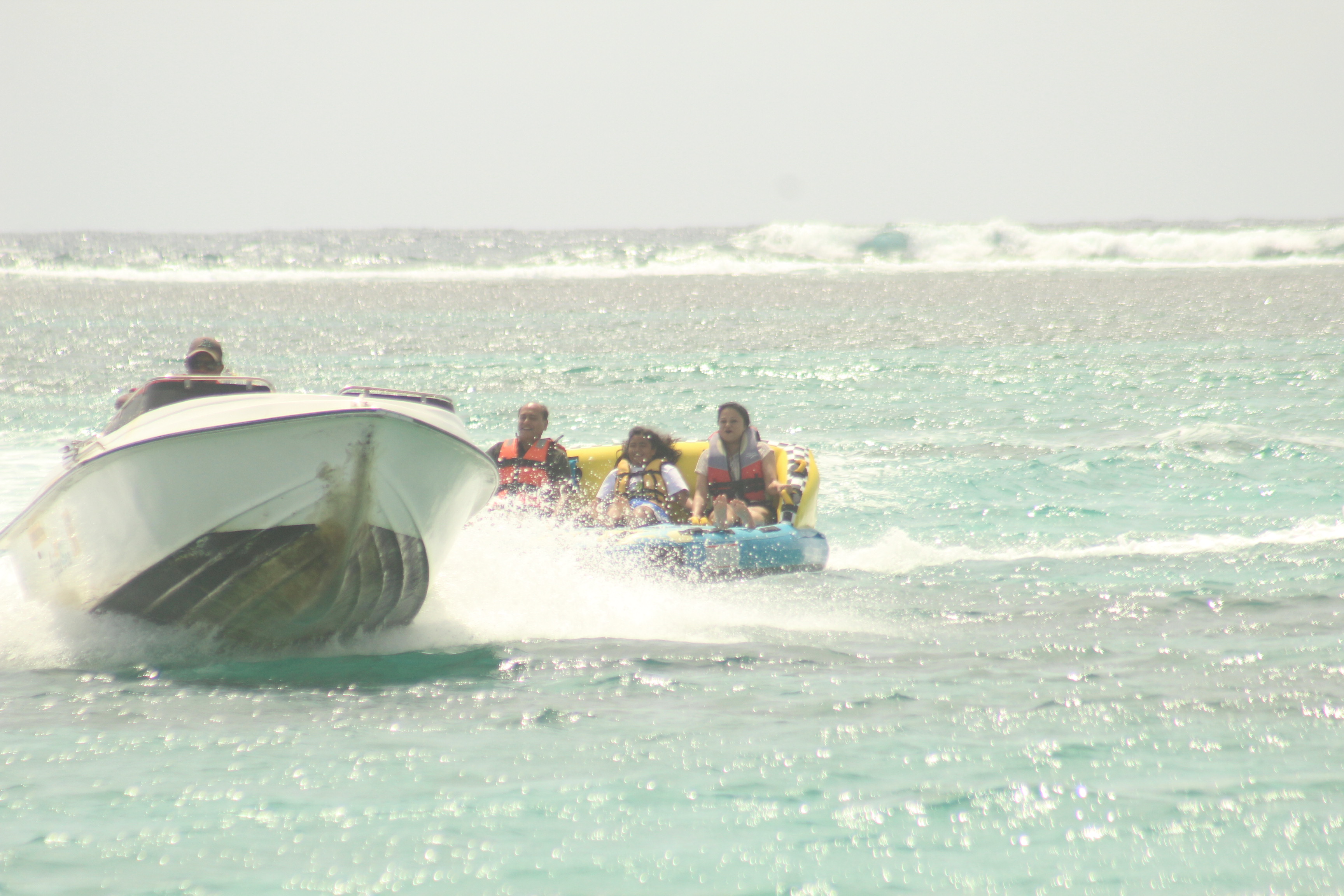 A speedboat pulls a group of people on a colorful inflatable tube in clear blue waters, with waves in the background.