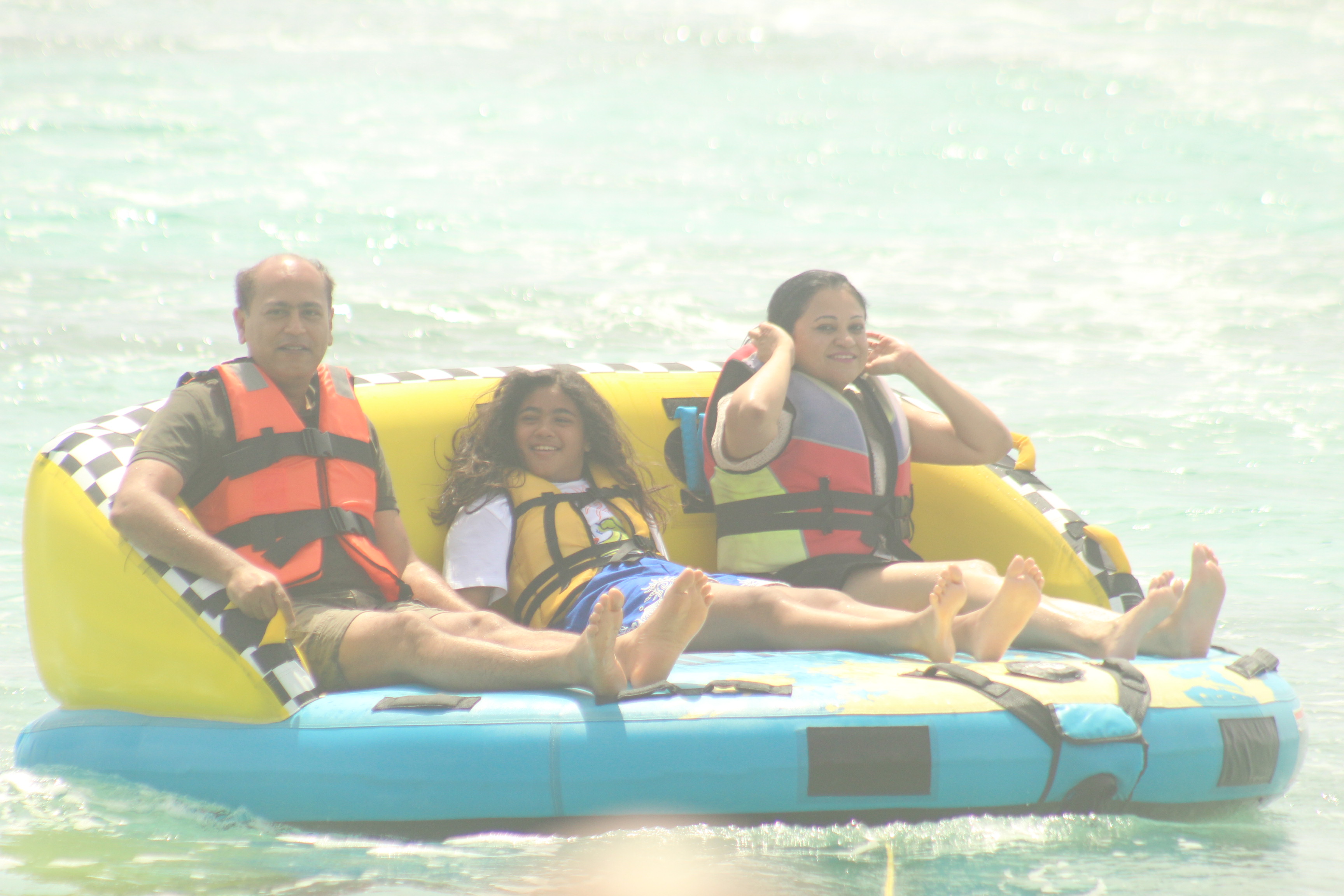 A family enjoying a ride on a yellow inflatable boat in the ocean, wearing life jackets. Two adults and a child are seated together, smiling and relaxed, with water sparkling in the background.